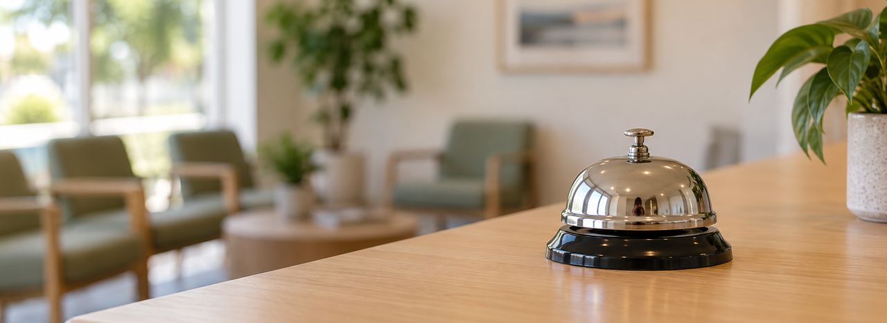 A chrome service bell on the timber reception counter of a calm, empty GP waiting room in morning light.